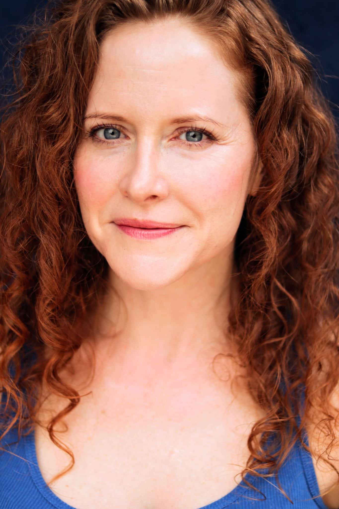 Woman with long, curly red hair and blue eyes, smiling softly, wearing a blue top. Blue background.