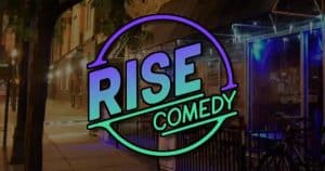 The image shows a street scene at night with RISE COMEDY in bold, colorful letters overlaid in a circular logo. Streetlights illuminate the sidewalk, and a nearby building has a glass facade with blue lights inside.
