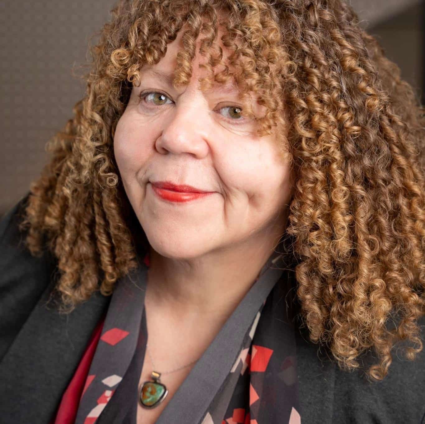 A person with curly, light brown hair smiles slightly at the camera. They wear a dark colored outfit with a patterned scarf and a green pendant necklace. The background is softly blurred.
