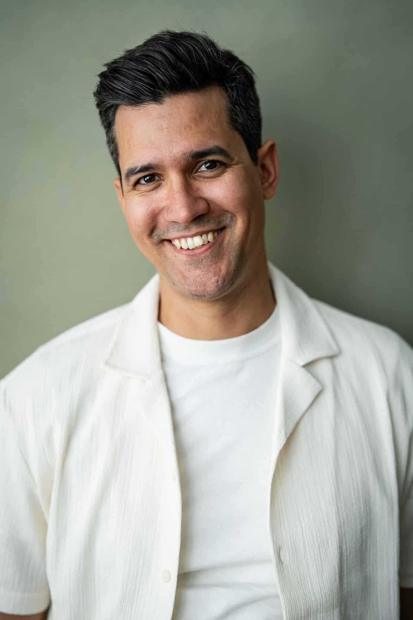 A man with short dark hair and medium skin tone smiles at the camera. He is wearing a white T-shirt and a light, short-sleeved shirt, standing against a plain, muted green background.
