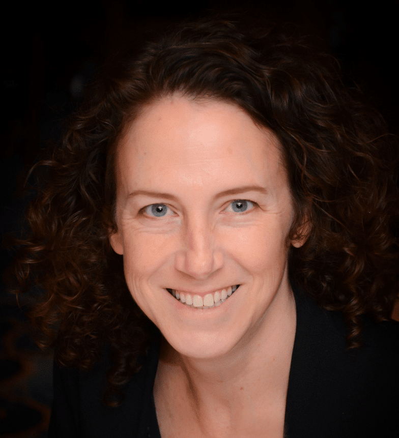 A woman with curly brown hair and blue eyes smiles at the camera. She is wearing a dark blazer and is pictured against a dark, softly blurred background.