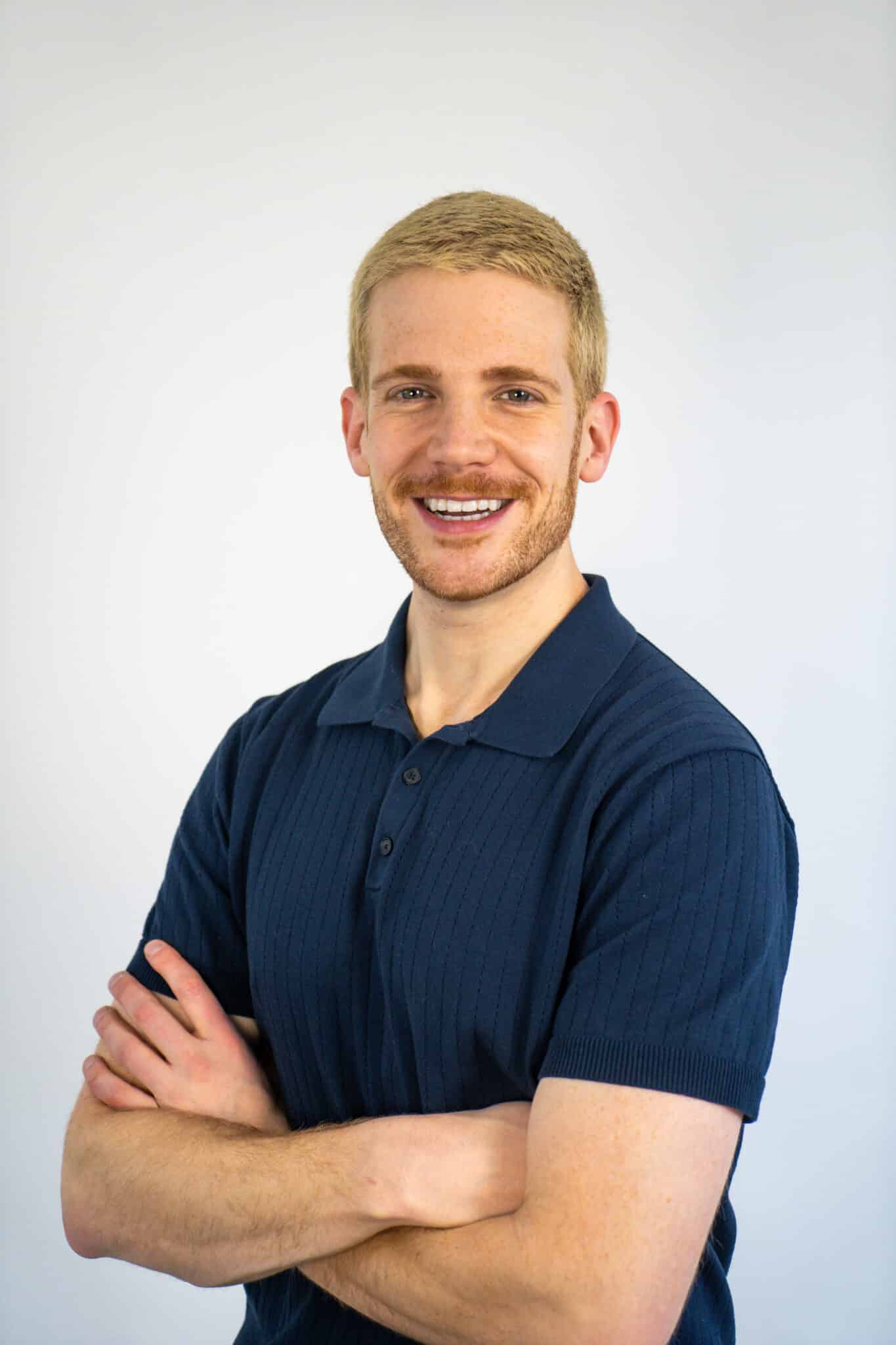 A smiling man with short blond hair and a trimmed beard stands against a plain light background, wearing a dark blue polo shirt and crossing his arms.