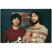Two young men with curly hair stand side by side indoors. One wears a maroon sweater with a graphic print, while the other wears a colorful patterned sweater and crosses his arms. A blurred sign is visible in the background.