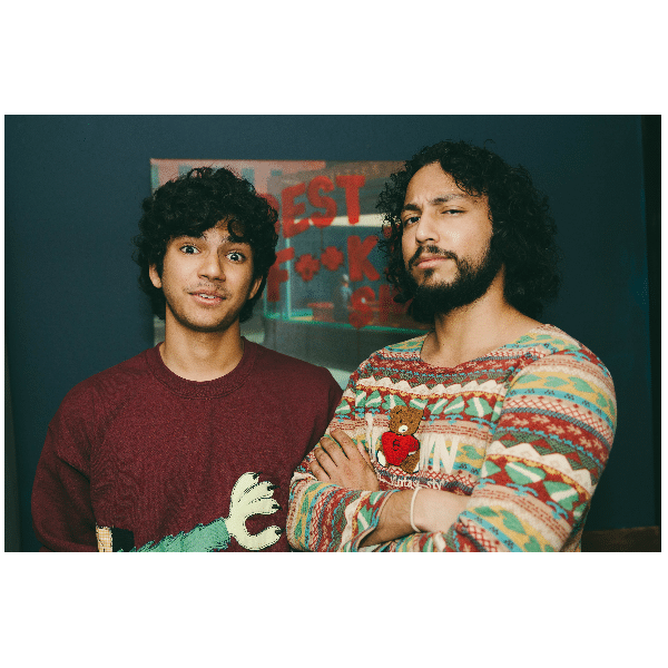 Two young men with curly hair stand side by side indoors. One wears a maroon sweater with a graphic print, while the other wears a colorful patterned sweater and crosses his arms. A blurred sign is visible in the background.