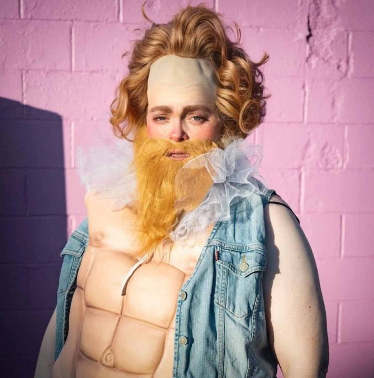 A person with a fake blond beard, a large forehead cap, and a ruffled white collar, wearing a denim vest and a fake muscular chest, stands in front of a pink brick wall in bright sunlight.