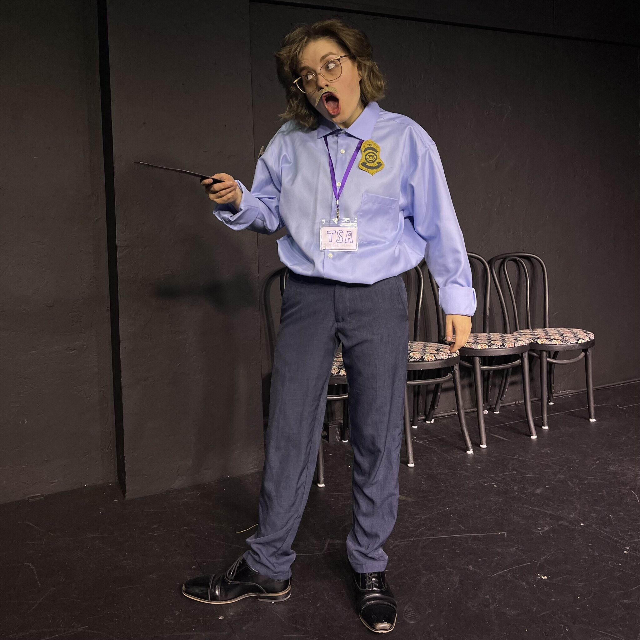 A person in a blue TSA shirt, holding a wand and making an expressive face, stands on a stage with black walls and several chairs lined up in the background.