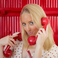 A woman with blonde hair holds two red retro telephone receivers to her ears, standing in front of a bright red background and wearing a white top with small heart patterns.