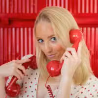 A woman with blonde hair holds two red retro telephone receivers to her ears, standing in front of a bright red background and wearing a white top with small heart patterns.