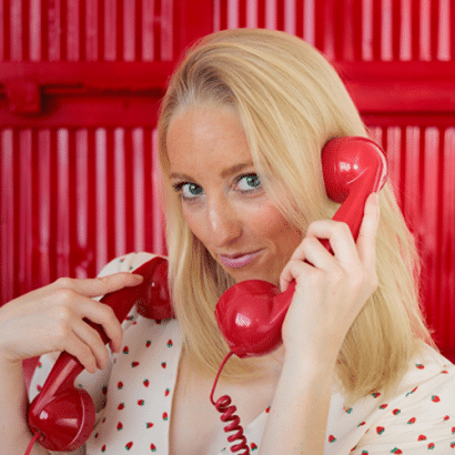 A woman with blonde hair holds two red retro telephone receivers to her ears, standing in front of a bright red background and wearing a white top with small heart patterns.