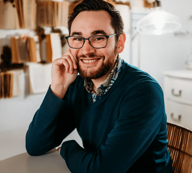 A man with glasses and a beard smiles while sitting at a desk, resting his head on his hand. Shelves of books and a lamp are visible in the background.